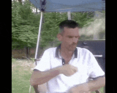 a man in a white shirt is sitting under an umbrella in front of a grill