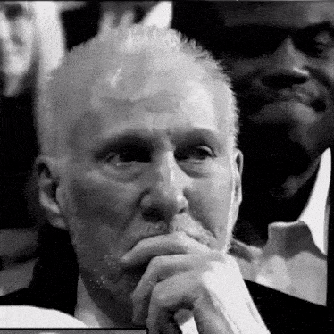 a black and white photo of an older man with a beard sitting in a crowd .