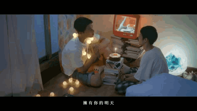 two men sit on the floor in front of a birthday cake with a candle