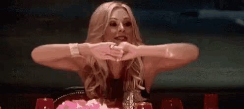 a woman is sitting at a table with her arms crossed and making a heart shape with her hands .