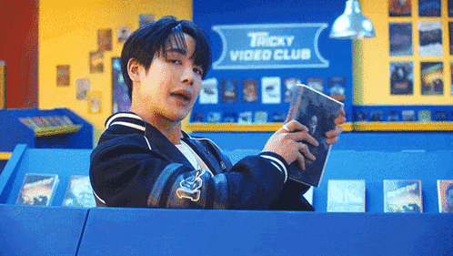a young man is sitting in a box holding a book in front of a sign that says tricky video club .