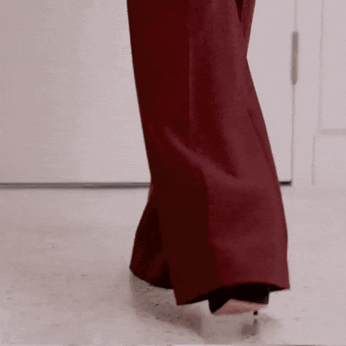 a woman wearing a pair of red pants and black heels is walking on a white floor .