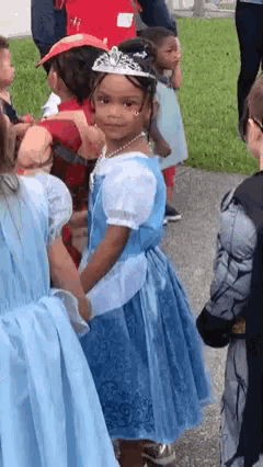 a little girl in a blue dress and tiara is standing in a crowd of children dressed as princesses .