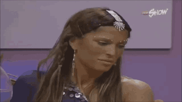 a woman wearing a headband and earrings is sitting in front of a white board with the word show on it .