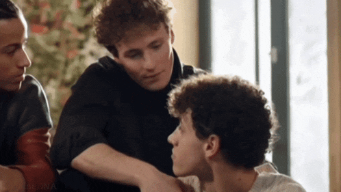 a group of young men are sitting around a table and one of them is touching another man 's neck .