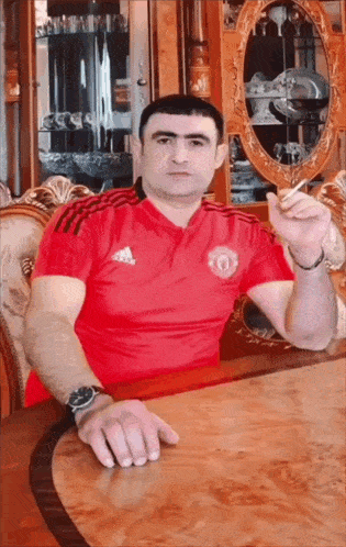 a man wearing a red adidas shirt is sitting at a table smoking a cigarette