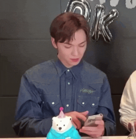 a man in a denim shirt is looking at his phone while sitting at a table .
