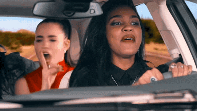 two women are sitting in a car with the letter d on the rearview mirror