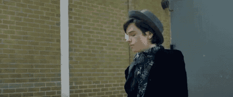 a young man wearing a hat and a scarf is standing in front of a brick wall looking at his watch .