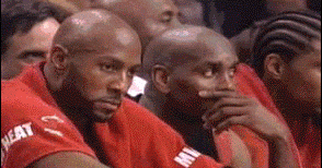 a group of basketball players are sitting in the stands watching a game and one of them is covering his mouth with his hand .