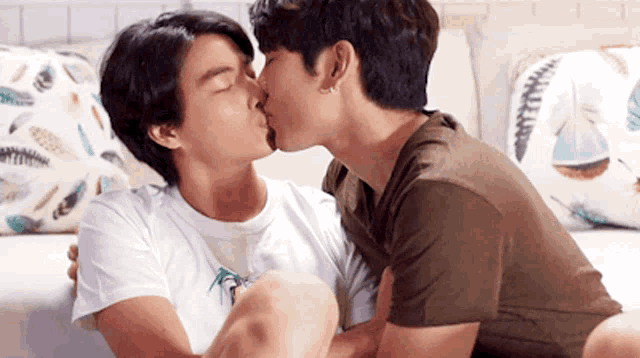 two men are kissing on a couch with pillows on it .
