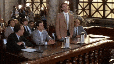 a man in a suit and tie stands in front of a group of men at a table