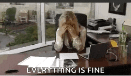 a woman is sitting at a desk with her head in her hands and the words everything is fine behind her .