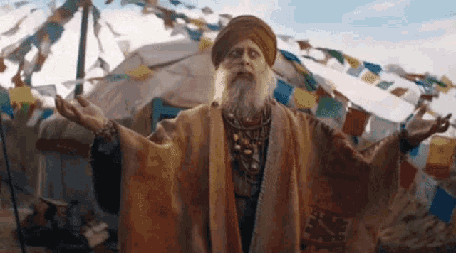 a man with a beard and turban is standing in front of a tent with flags flying in the wind .