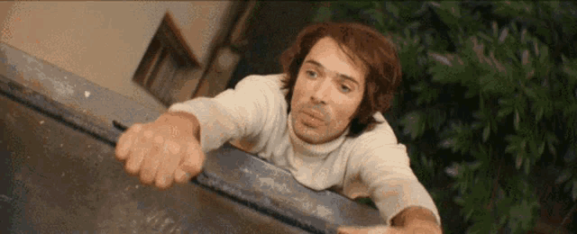 a man in a white sweater is leaning over a metal fence .