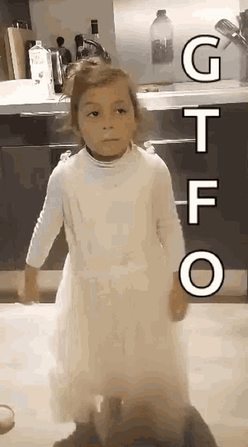 a little girl in a white dress is standing in a kitchen with the letters gtfo written on the wall behind her .