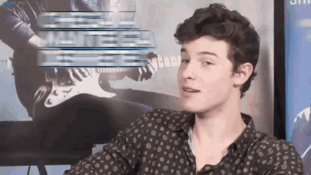 a man in a polka dot shirt is sitting in front of a poster of a man playing an electric guitar