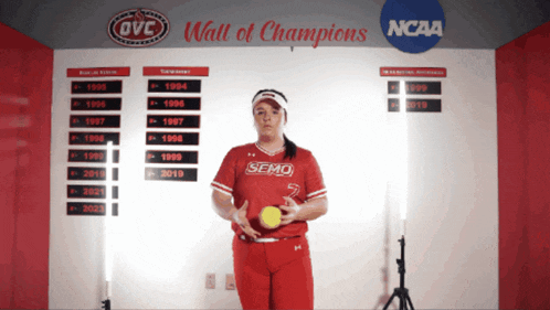 a woman holding a softball in front of a wall of champions sponsored by ncaa
