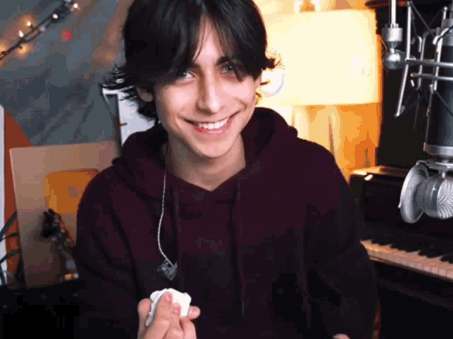 a young man in a maroon hoodie smiles in front of a piano