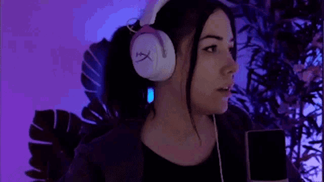 a woman wearing headphones is pointing at the camera while holding a cell phone .