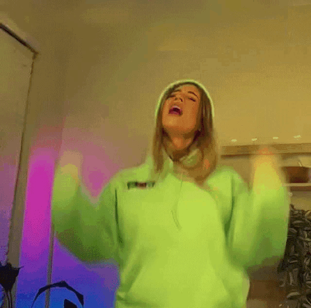 a woman in a neon green hoodie is dancing in a room with her arms in the air .