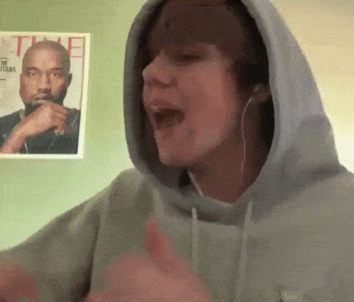 a man wearing a hoodie and headphones is singing in front of a picture of kanye west .