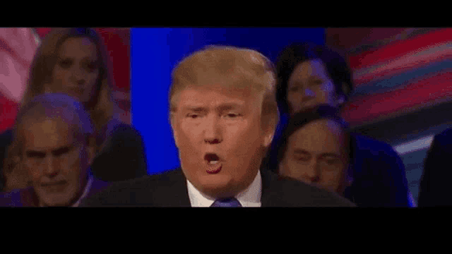 donald trump is giving a speech in front of a crowd of people while wearing a suit and tie .