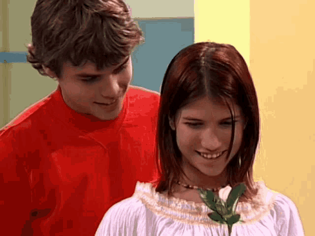 a man in a red shirt stands next to a woman in a white dress holding a flower