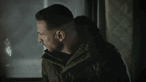 a man with a beard is wearing a jacket and looking out a window