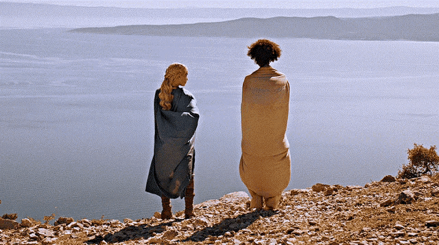 two people wrapped in blankets standing on a rocky cliff overlooking a body of water