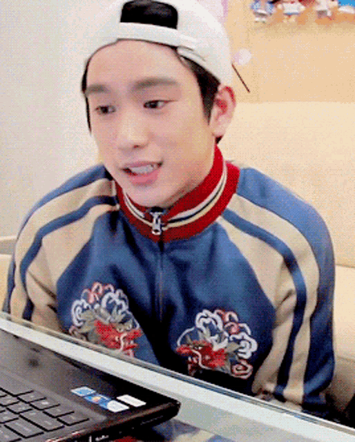 a young man wearing a baseball cap and a blue jacket sits in front of a laptop computer