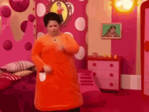 a woman in an orange dress is dancing in a pink room with a picture of a woman on the wall .