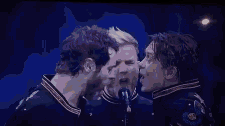 three men are singing into microphones on a stage and one of them is kissing the other .