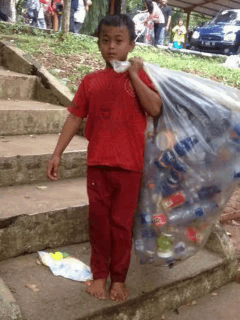 a young boy in a red shirt is carrying a bag full of plastic bottles and cans