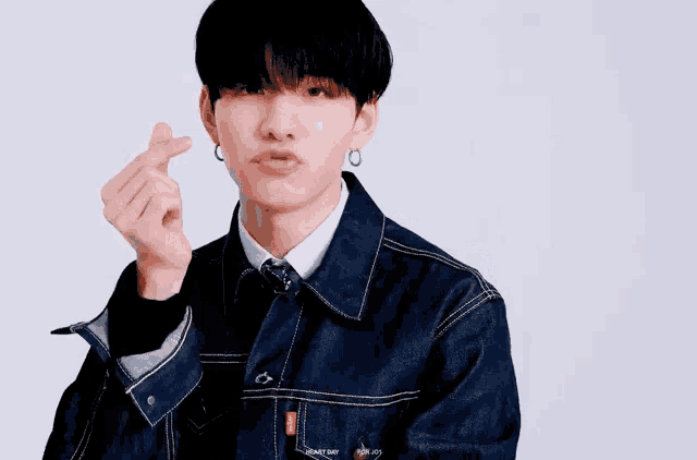 a young man in a denim jacket and tie is making a heart with his hands .
