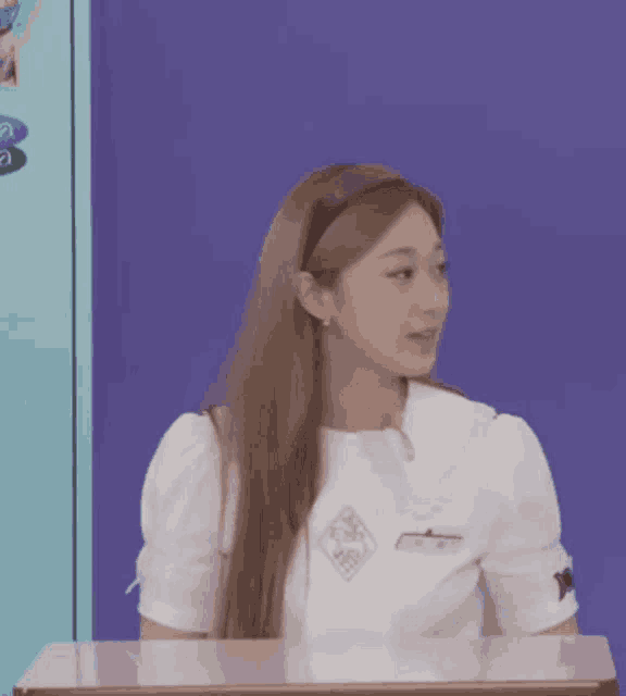 a woman in a white shirt is sitting at a desk in front of a purple wall