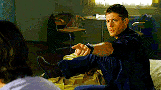 a man is sitting on a bed with his legs crossed and pointing at another man .