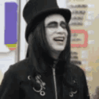 a man with long black hair and white face paint is wearing a top hat and laughing .