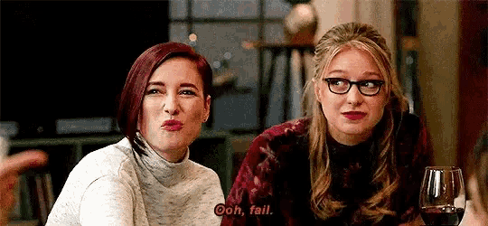 two women are sitting next to each other at a table with a glass of wine and making funny faces .