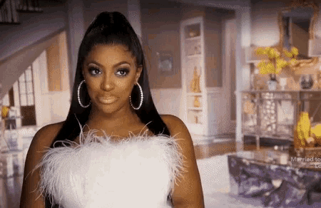 a woman wearing a white feathered top and hoop earrings stands in a living room