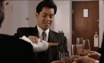 a man in a suit and tie is sitting at a table with glasses of wine and a cigar .