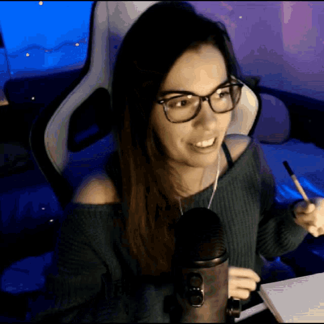 a woman wearing glasses sits in front of a microphone holding a pen