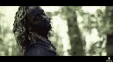 a man with long curly hair is standing in a forest .