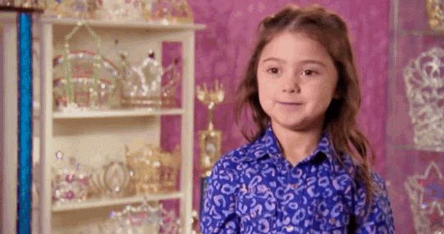 a little girl in a blue shirt is standing in front of a shelf filled with crowns .