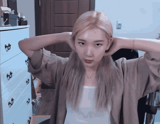 a woman with blonde hair holds her hands behind her head in front of a dresser