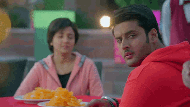 a man in a red hoodie looks at a woman sitting at a table with plates of food