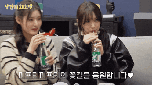 two girls are sitting on a couch drinking soda from bottles