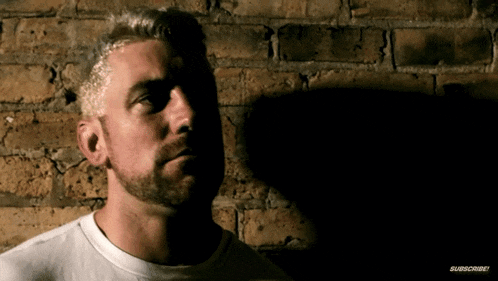a man with a beard is standing in front of a brick wall with a subscribe button on the bottom right