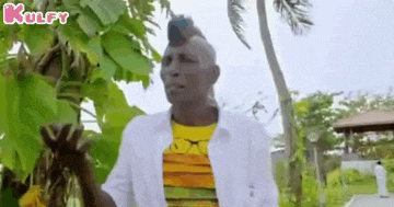 a man with a mohawk is standing in front of a palm tree holding a banana .