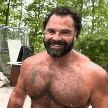 a shirtless man with a beard is smiling while standing in front of a fence .